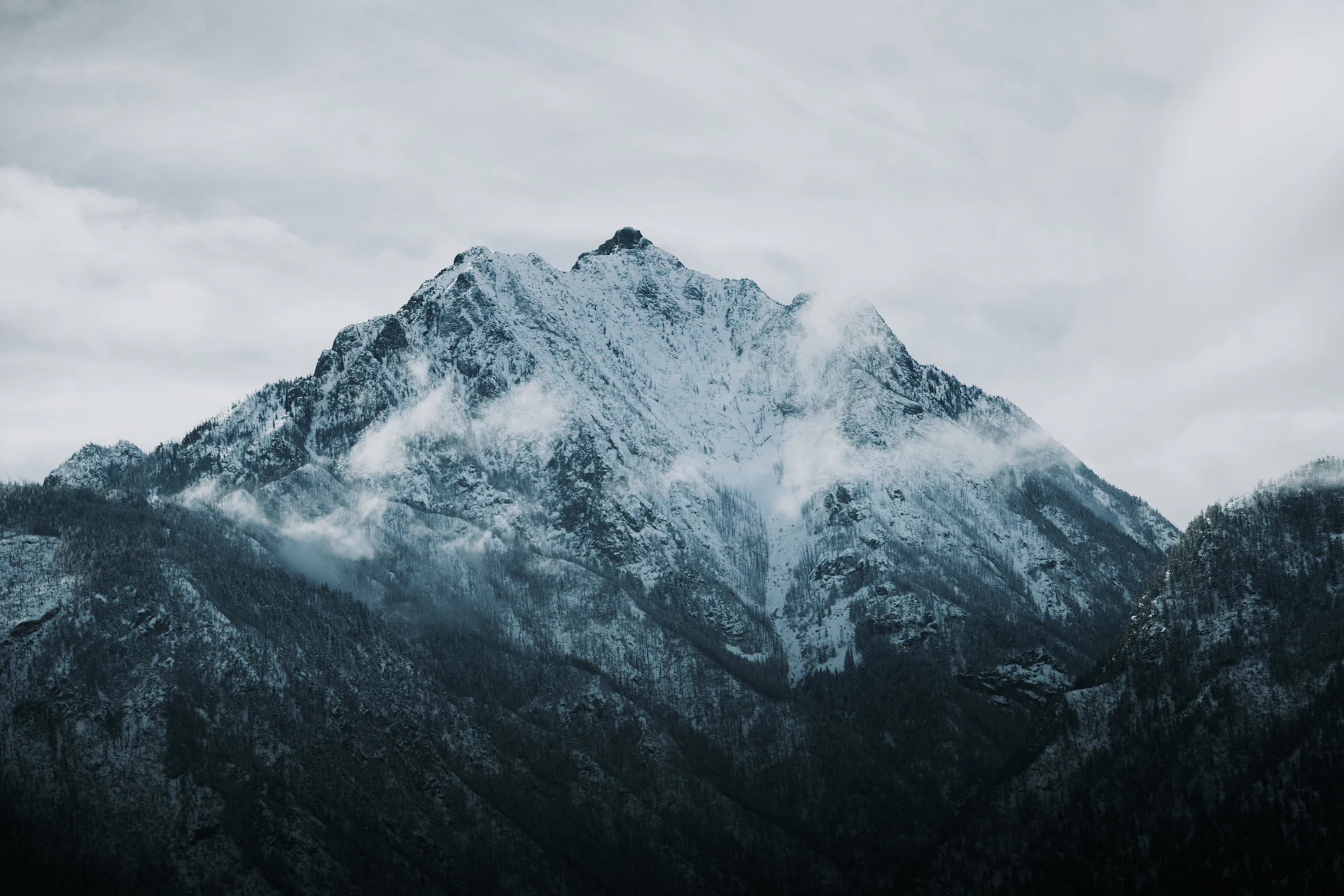 Snow-covered mountain peak under a pale overcast sky, above dark rocky slopes
